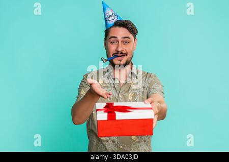 Un uomo di mezza età che indossa un cappello festoso sorride calorosamente, fa felicemente un regalo di compleanno, celebrando chiaramente la generosità e la gentilezza. Ragazzo adulto isolato su sfondo blu che si gode una festa gioiosa Foto Stock