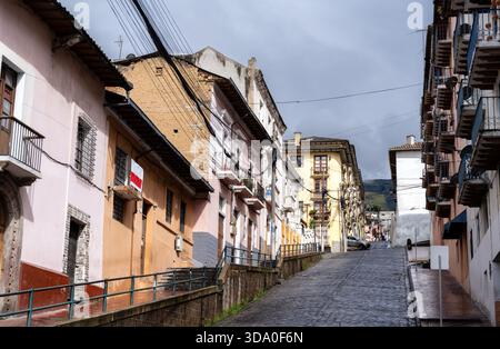 Ripida strada acciottolata fiancheggiata da case coloniali colorate a Quito, Ecuador. L'architettura storica presenta balconi e pareti color pastello nel sito patrimonio dell'umanità dell'UNESCO vicino alla Basilica Foto Stock