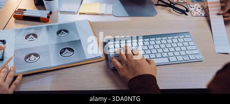 Un'area di lavoro creativa vivace con un computer, strumenti di progettazione, campioni di colore e mani di designer che collaborano a un progetto di branding Foto Stock