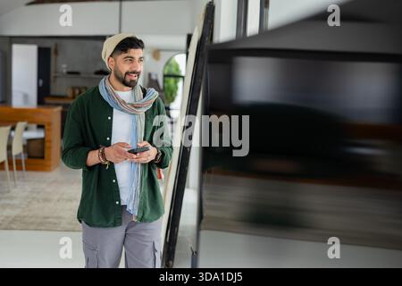 Uomo indiano che tiene in mano uno smartphone che guarda il display accanto agli sgabelli della cucina dell'isola nel loft, spazio fotocopie Foto Stock