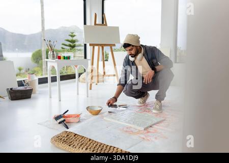 Pittore indiano accovacciato su stoffa dipinta in studio dipingendo grandi tele vicino alle finestre Foto Stock