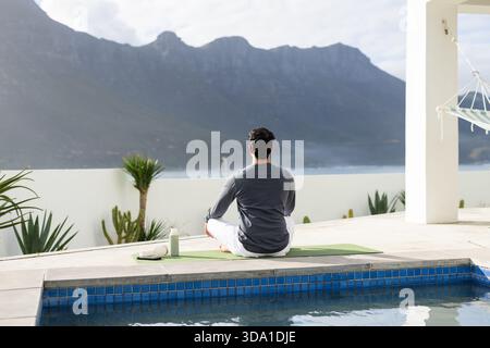 Uomo indiano che meditava sul tappetino verde per yoga sulla terrazza a bordo piscina vicino alla piscina con piastrelle blu e bottiglia d'acqua Foto Stock