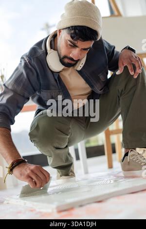 Uomo indiano accovacciato in uno studio d'arte che diffonde medio bianco su grande tela con cazzuola vicino al cavalletto Foto Stock
