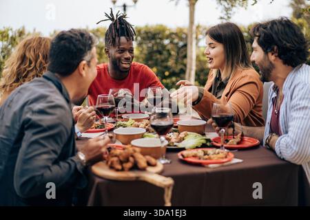 Gruppo di amici seduti al tavolo, a parlare e a gustare del vino durante una rilassante cena all'aperto. Atmosfera calda, emozioni reali, persone diverse, cibo Foto Stock