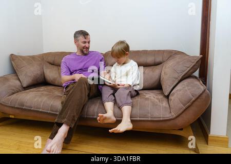Un padre e un figlio siedono a piedi nudi su un divano marrone a leggere un libro. L'uomo sorride al ragazzo, che è concentrato sulla storia. Un momento informale in famiglia a casa. Foto Stock