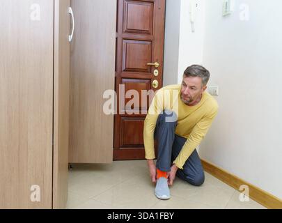 Un uomo di mezza età con una camicia gialla e pantaloni grigi si inginocchia su un pavimento piastrellato in un ingresso di casa, indossando le sue scarpe slip-on grigie vicino alla porta d'ingresso. Foto Stock