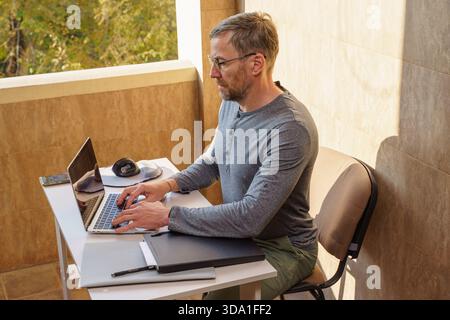 Un uomo di mezza età con gli occhiali lavora a distanza da un balcone soleggiato. Si siede a una scrivania bianca, scrivendo su un notebook, con un tablet grafico nelle vicinanze Foto Stock