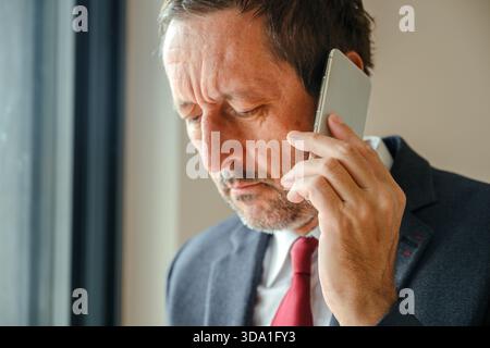 Un uomo d'affari preoccupato in tuta formale che parla al telefono cellulare vicino alla finestra dell'ufficio, ha sottolineato la comunicazione aziendale. Messa a fuoco selettiva. Foto Stock