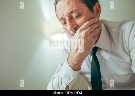Stanco uomo d'affari che sbadiglia con la mano che copre la bocca, mostrando stanchezza e mancanza di sonno. Messa a fuoco selettiva. Foto Stock