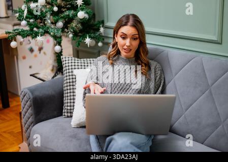 Seduto comodamente in un comodo maglione, la donna penosa sta usando il suo computer portatile per le videochiamate. Comunica online. Foto Stock