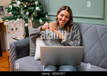 Una donna sorridente si rilassa su un divano con una bevanda calda in mano, lavorando sul suo portatile mentre un albero di Natale decorato si illumina sullo sfondo. Foto Stock