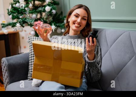 Una donna felice si siede comodamente su un divano, reggendo profumi e consegnando il pacco senza confezione. Albero di Natale decorato sullo sfondo. Foto Stock