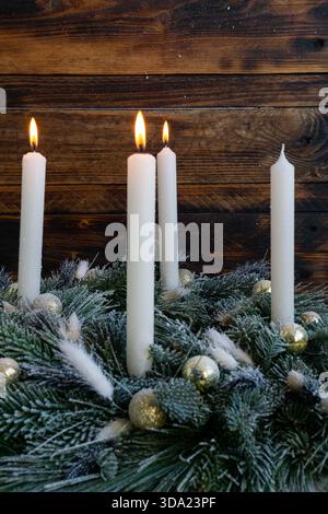 Tre alte candele bianche risplendono calorosamente sopra una corona sempreverde smerigliata decorata con ornamenti dorati. Foto Stock