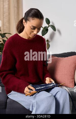 Una giovane donna afroamericana ama lavorare a maglia mentre si rilassa sul suo divano in un appartamento moderno. Foto Stock
