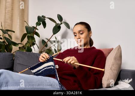Una giovane donna afro-americana si diverte a lavorare a maglia in un accogliente maglione invernale a casa. Foto Stock