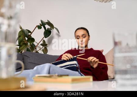 Una giovane donna afroamericana ama lavorare a maglia nel suo moderno appartamento durante l'inverno. Foto Stock
