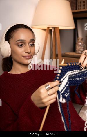 Una giovane donna afroamericana ama lavorare a maglia indossando le cuffie nel suo elegante appartamento. Foto Stock