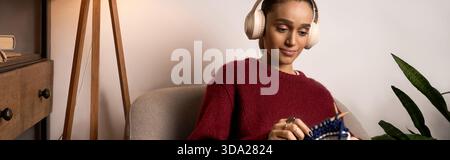 La giovane donna afroamericana gode di un momento di pace lavorando a maglia nel suo accogliente appartamento, banner Foto Stock