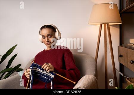 La giovane donna afroamericana ama trascorrere un pomeriggio rilassante a maglia a casa, indossando un maglione marrone. Foto Stock