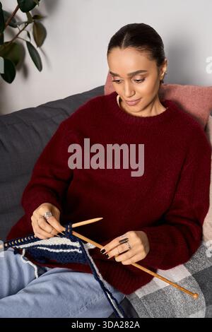 Una giovane donna ama lavorare a maglia nel suo elegante appartamento indossando un comodo maglione marrone. Foto Stock