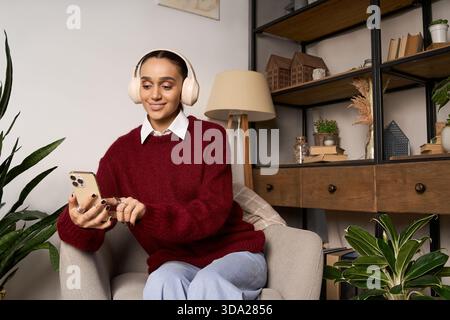 Una giovane donna sorride mentre usa il telefono, godendosi il calore del suo maglione a casa. Foto Stock