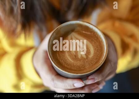 Donna che tiene a mano una tazza calda di caffè con latte art a forma di cuore. Vista dall'alto della donna che si gode momenti intimi con una bevanda calda, si sente rilassata e confortevole Foto Stock