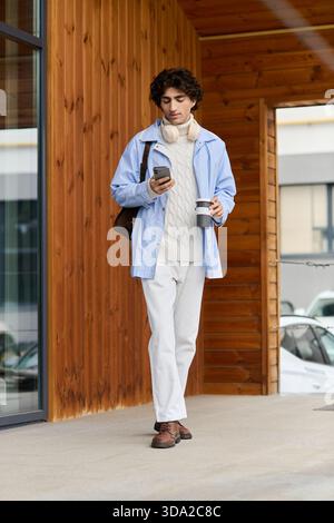 Un giovane con i capelli ricci passeggia per la città, sorseggiando un caffè e controllando il telefono. Foto Stock