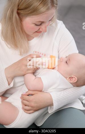 La mamma felice tiene il suo bambino appena nato tra le braccia e lo nutre dal biberon, mostrando attenzione al bambino. Concetto di allattamento al neonato, alternativa all'allattamento al seno, Foto Stock