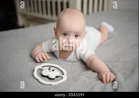 Un bambino appena nato carino con guance paffute giace sullo stomaco e gioca con il giocattolo sul letto. Neonati, prodotti per bambini e giocattoli Foto Stock
