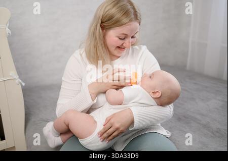 Una donna felice tiene il bambino tra le braccia e lo nutre dal biberon, mostrando la cura per il bambino. Assistenza sanitaria, concetto di allattamento al neonato, alternativa al seno Foto Stock