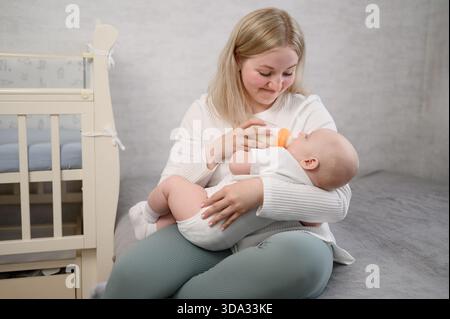 La mamma tiene il bambino in braccio, seduto sul letto accanto alla culla e nutre il neonato con il latte in polvere dal biberon, mostrando preoccupazione per il bambino. Assistenza sanitaria, neonato Foto Stock