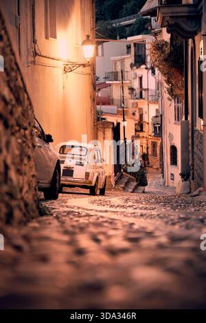 Calda strada di ciottoli serale in una città vecchia mediterranea con una piccola auto d'epoca e luci calde. Foto Stock