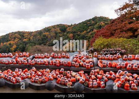 Le bambole Daruma sono collocate su un tetto di un tempio. La scena mostra alberi colorati sullo sfondo, a indicare l'autunno. Le nuvole riempiono il cielo. La gente visita Foto Stock