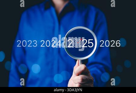 Concentrarsi sugli obiettivi aziendali del 2025 e sulla pianificazione futura. Una persona con una lente d'ingrandimento che evidenzia 2025 con un'icona di obiettivo, obiettivi futuri, strategia, Foto Stock