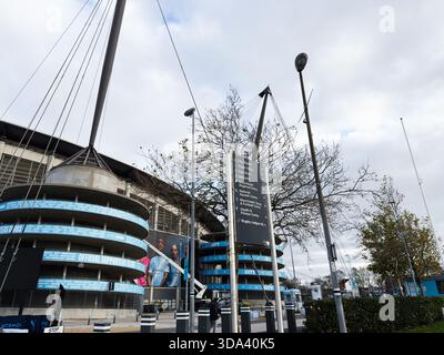 Manchester, Regno Unito - 16 novembre 2025: Vista esterna del City of Manchester Stadium con striscioni blu e segnaletica urbana. Foto Stock