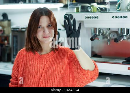 La donna sorridente in maglione arancione solleva la mano con un guanto nero, mostrando quattro dita vicino alla macchina per espresso. Foto Stock