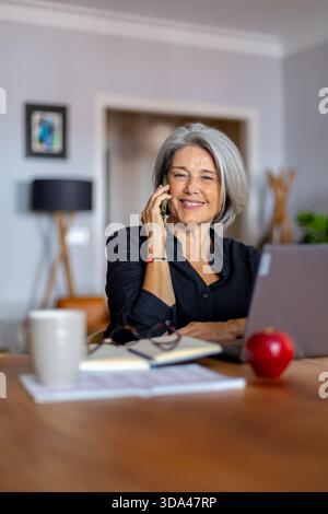Donna d'affari dai capelli grigi che parla con lo smartphone e lavora con il computer portatile in ufficio domestico, mentre lavora in remoto e lavora in orari flessibili Foto Stock
