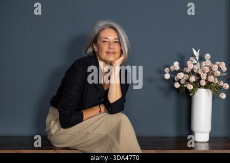 Ritratto di una donna d'affari sorridente e matura seduta su una panchina di legno con la mano sul mento, capelli grigi e fiori in vaso Foto Stock