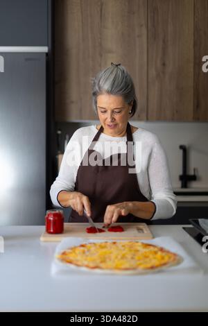 Donna anziana che taglia la paprika rossa su un asse di legno, preparando condimento fresco per una pizza fatta in casa su un bancone della cucina Foto Stock