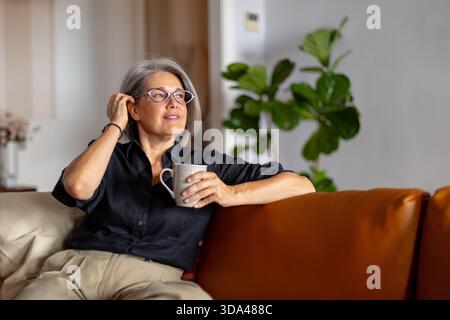Donna matura dai capelli grigi con occhiali da vista che si gode una tazza di caffè o tè mentre si siede su un comodo divano nel suo moderno soggiorno Foto Stock
