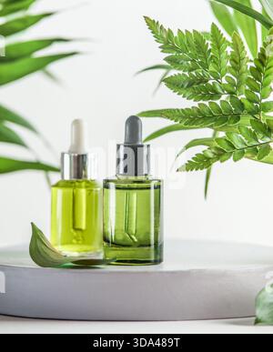 Flaconi per siero verde o olio essenziale con pipetta su podio con foglie tropicali su sfondo bianco. Cura della pelle moderna e sana con bellezza naturale pro Foto Stock