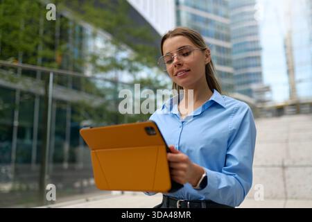 Giovane donna in camicia blu e occhiali sorridente mentre lavora su un tablet con una custodia arancione all'aperto, edifici in vetro urbano sullo sfondo. Edificio moderno Foto Stock