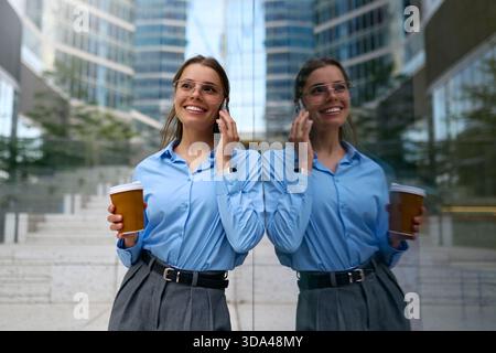 Giovane donna d'affari felice con camicia blu e bicchieri che reggono il caffè, parlano sullo smartphone, si trova accanto a una parete a specchio con riflessi nell'architettura moderna Foto Stock