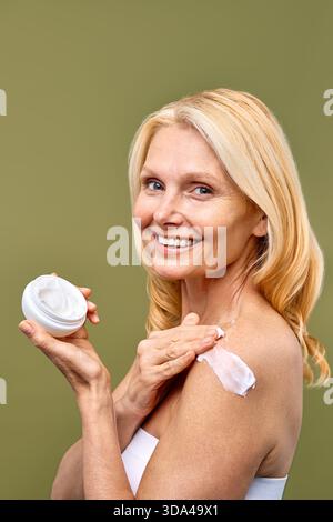 Un'immagine di una donna matura sorridente mentre si applica crema idratante sulla spalla incarna una sana routine di cura della pelle e antiaging cura per il radiano Foto Stock