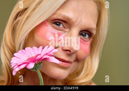 Una donna matura con i capelli biondi tiene un fiore rosa, mostrando la sua bellezza naturale e la cura di sé con cerotti per la cura della pelle sotto gli occhi. Cosmetologia, spa a Foto Stock