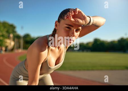 Primo piano di una giovane donna stanca che pulisce la fronte dopo aver corso in pista. Concetto di sforzo, recupero e perseveranza nell'allenamento di fitness. Foto Stock