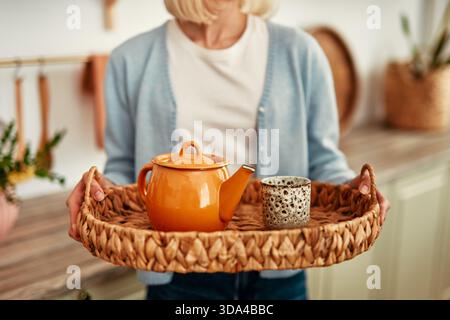 Una donna in un cardigan azzurro, in un'accogliente cucina, presenta una teiera arancione e una tazza in ceramica a motivi geometrici su un vassoio intrecciato, che irradia fascino e calore Foto Stock