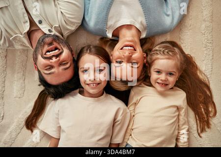 Una famiglia felice di quattro persone sdraiata sul tappeto, sorridente, coccolata e giocata felicemente. I genitori e i bambini si divertono a trascorrere un po' di tempo insieme, creando un caldo memor Foto Stock