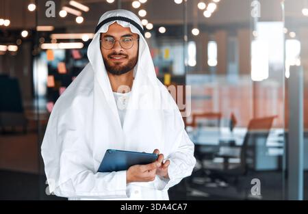 Giovane uomo d'affari del Medio Oriente in abiti tradizionali degli Emirati che utilizza un tablet in ufficio. Team aziendale che lavorano e brainstorming in UA Foto Stock