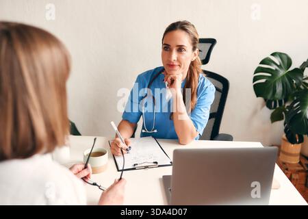 Medico donna in uniforme medica con stetoscopio seduto a tavolo con laptop prendendo appunti e parlando con il paziente nello studio medico in ospedale. Ciao Foto Stock